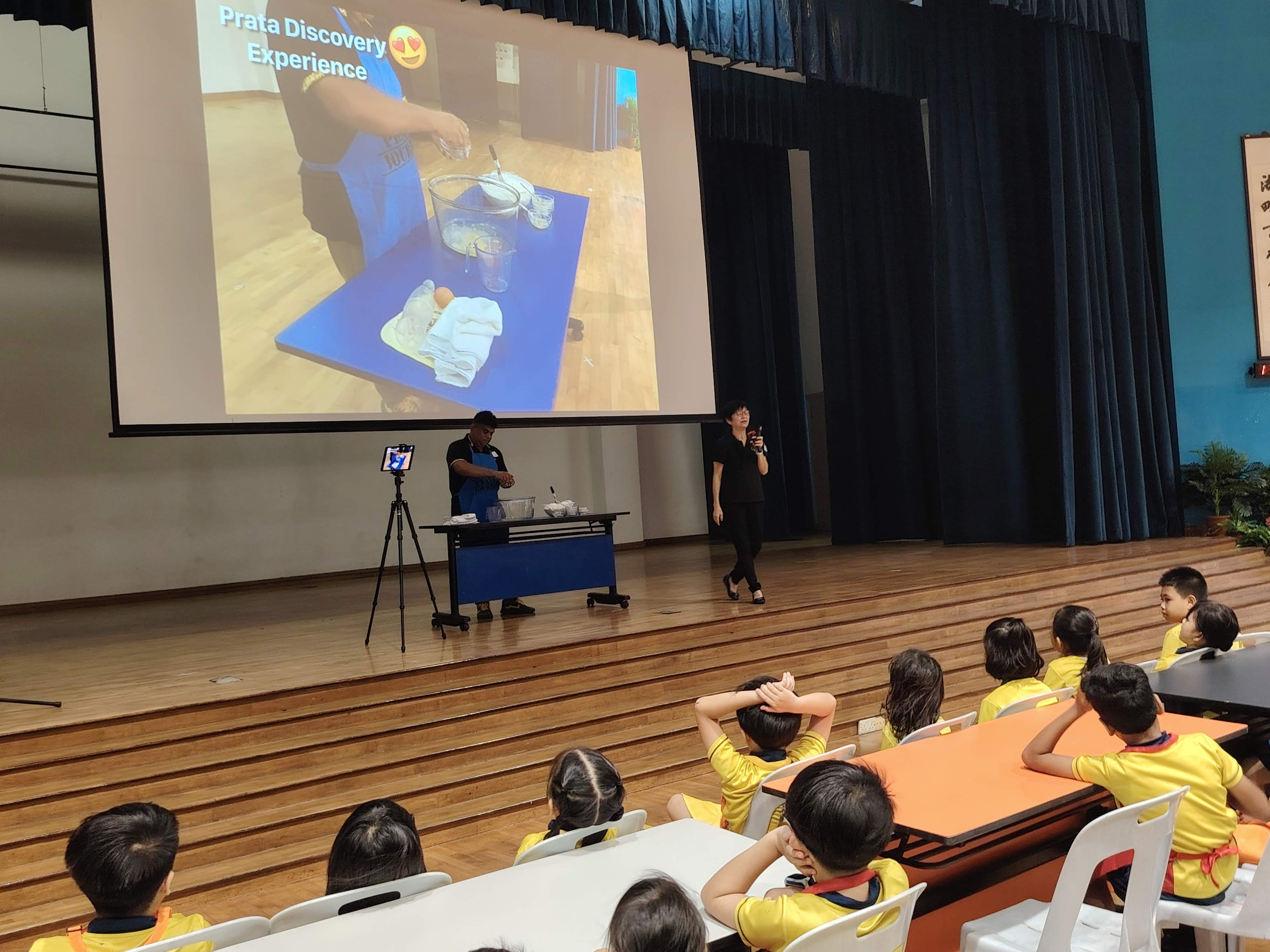 A chef on stage doing a live demonstration of making roti prata dough via livestreaming on the screen to the students watching from the the audience